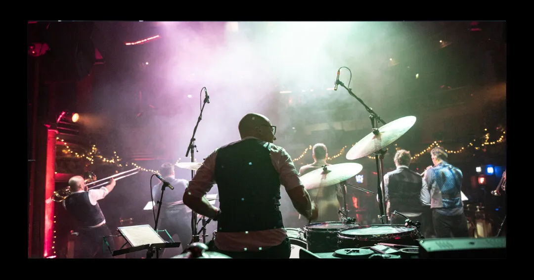 Rock Star Brass on stage seen from behind the drummer, mid-performance under smoke and stage lights.